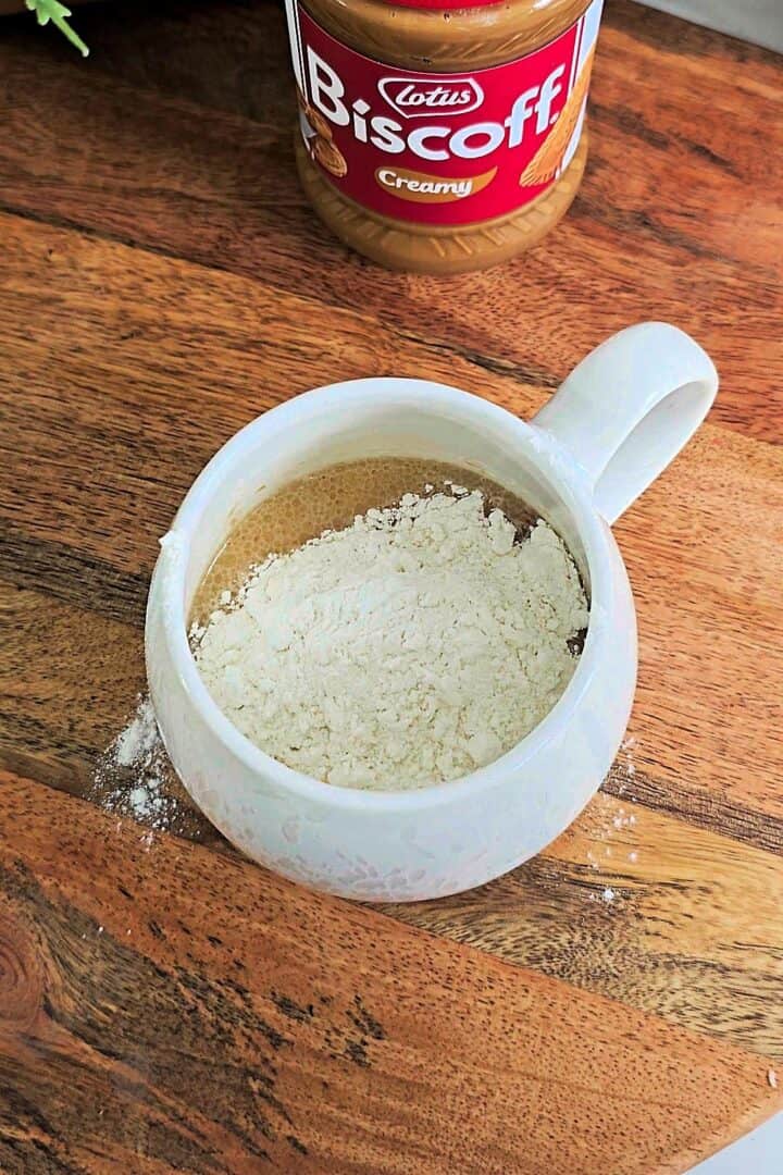 All ingredients including flour added to a white mug on a wooden board before mixing.