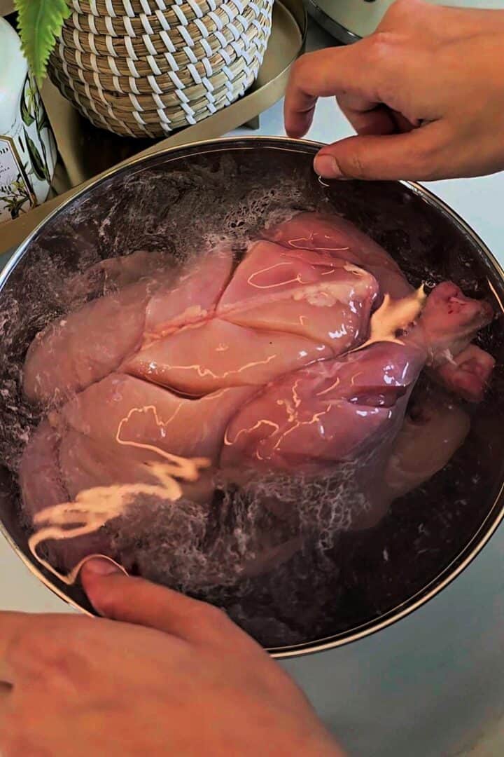 Raw whole chicken submerged in salted water brine in a metal bowl.