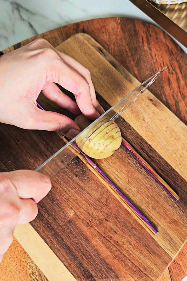 Potato placed between chopsticks while being sliced into even cuts on a wooden board.