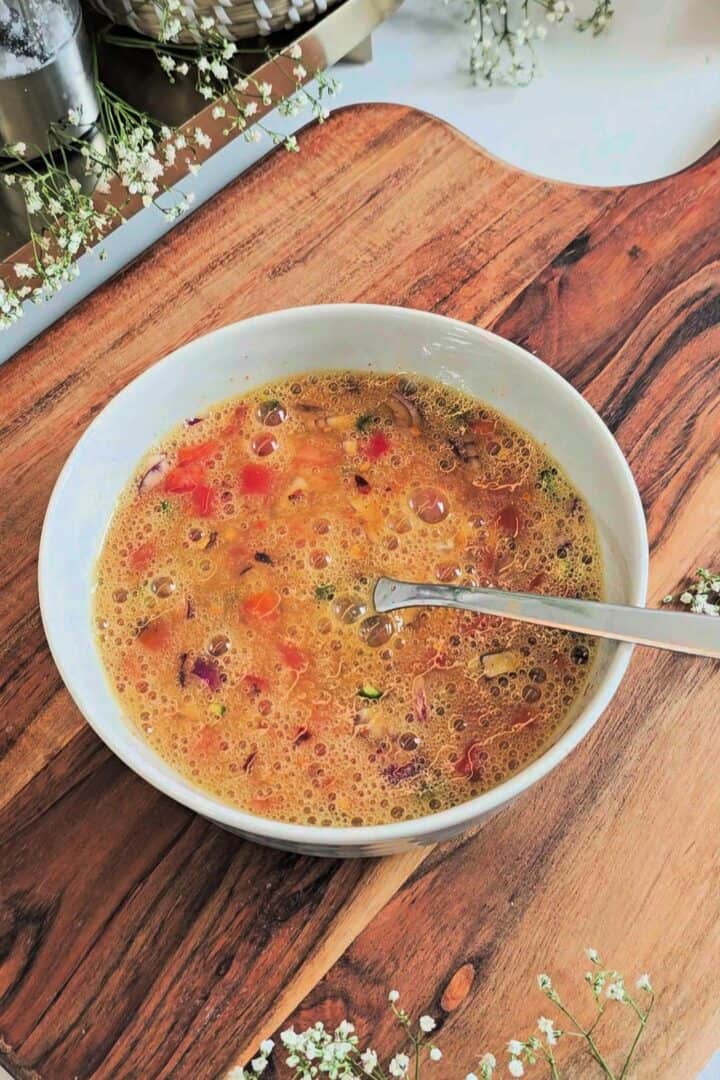 Bowl of whisked egg mixture with chopped onions, tomatoes, and herbs on a wooden board with a spoon.