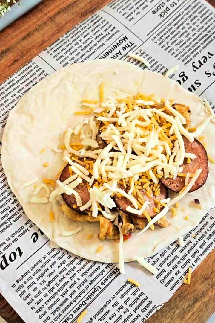 Paratha topped with scrambled eggs, sliced bologna, and shredded cheese on parchment paper.