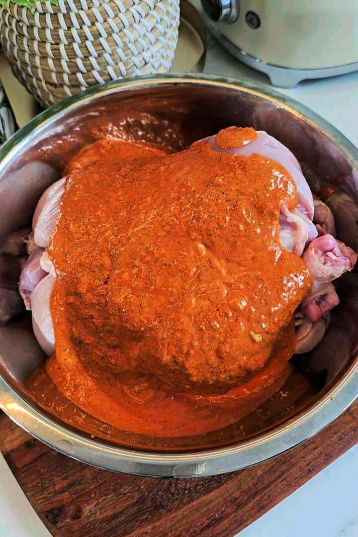Whole chicken coated in thick orange marinade in a metal bowl.