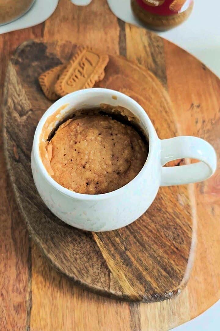 Freshly microwaved Biscoff mug cake in a white mug on a wooden board.