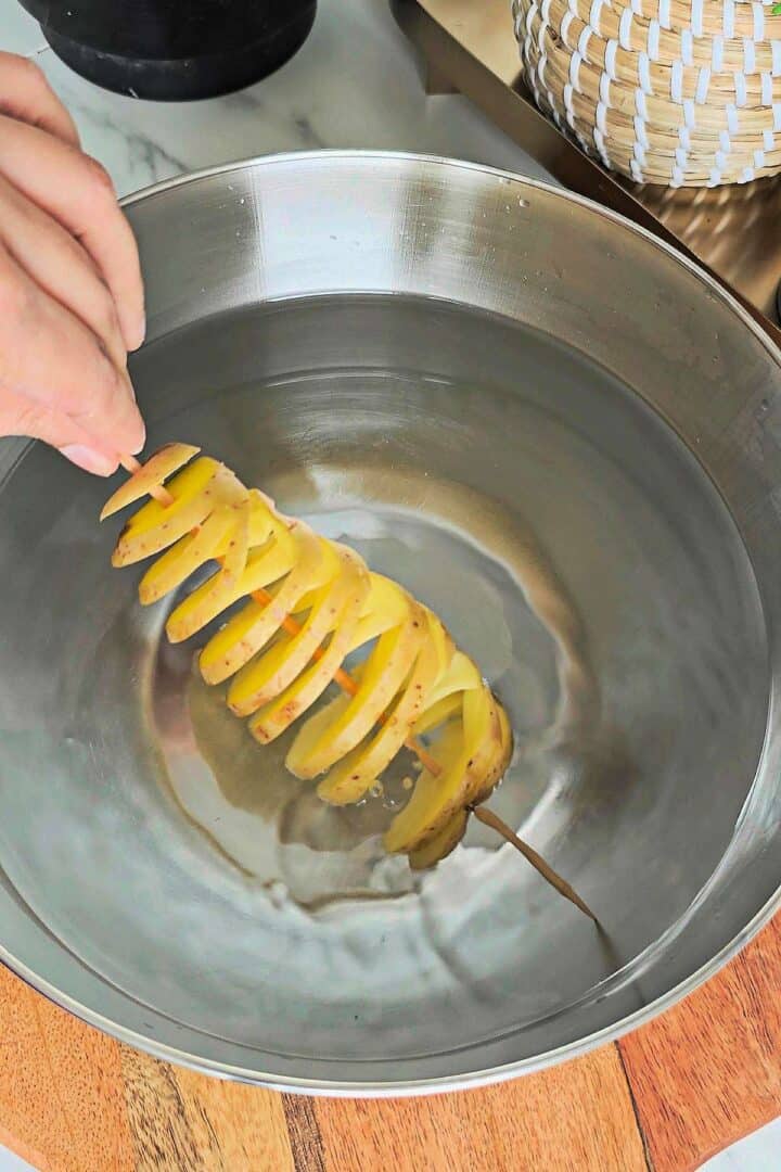 Spiral potato on a skewer being dipped into a bowl of water for soaking.
