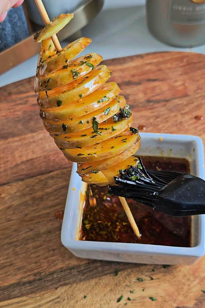 Spiral potato on a skewer being brushed with seasoned oil over a bowl.