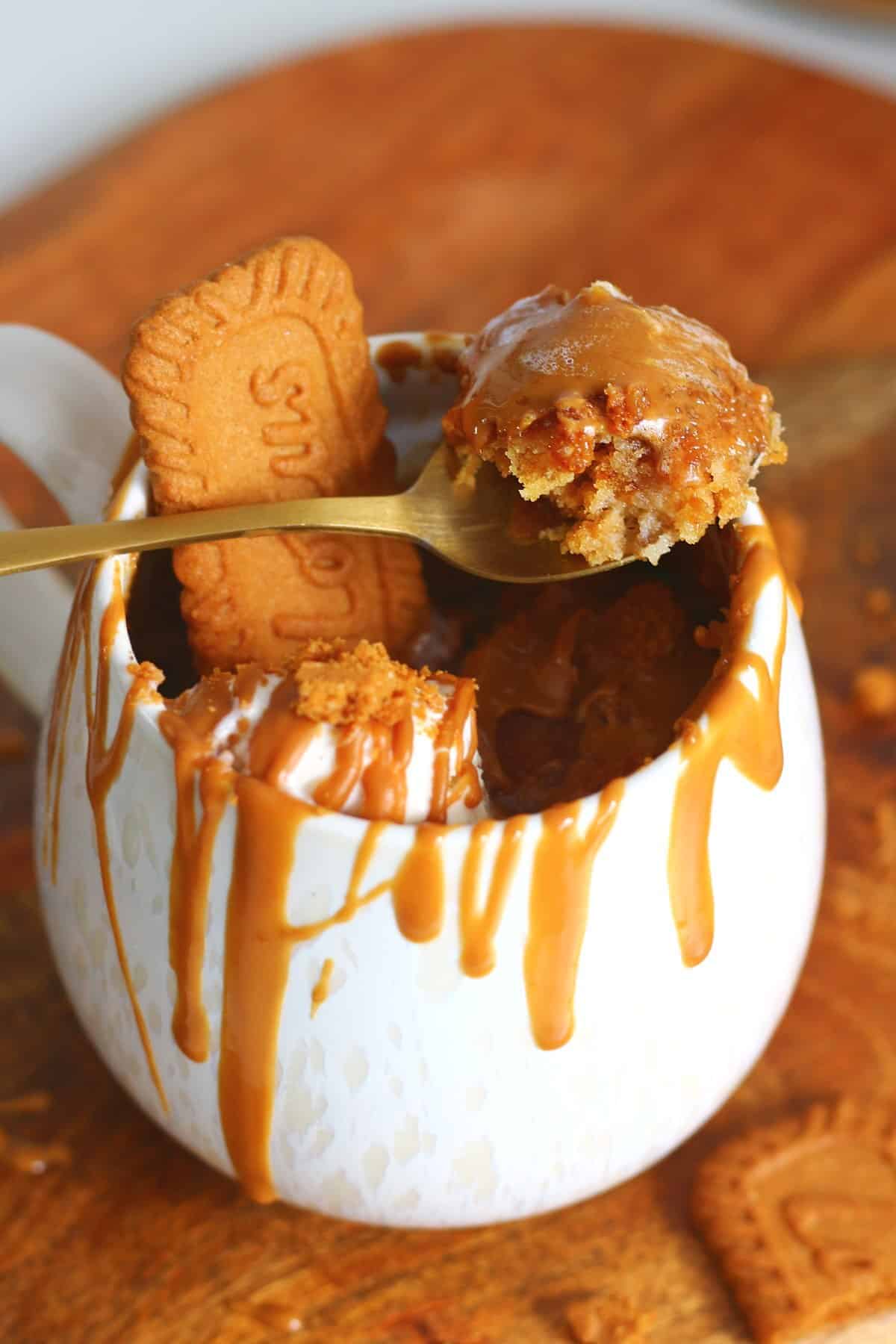 Spoon lifting a bite of Biscoff mug cake with melted Biscoff and ice cream from a white mug.