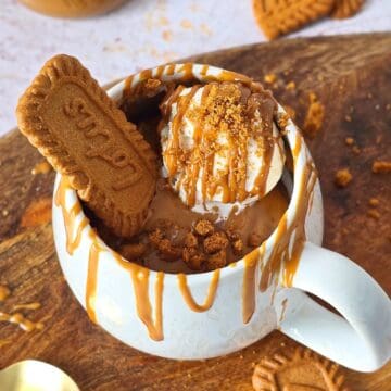 Biscoff mug cake topped with ice cream, Biscoff drizzle, and a whole Biscoff cookie on a wooden board.