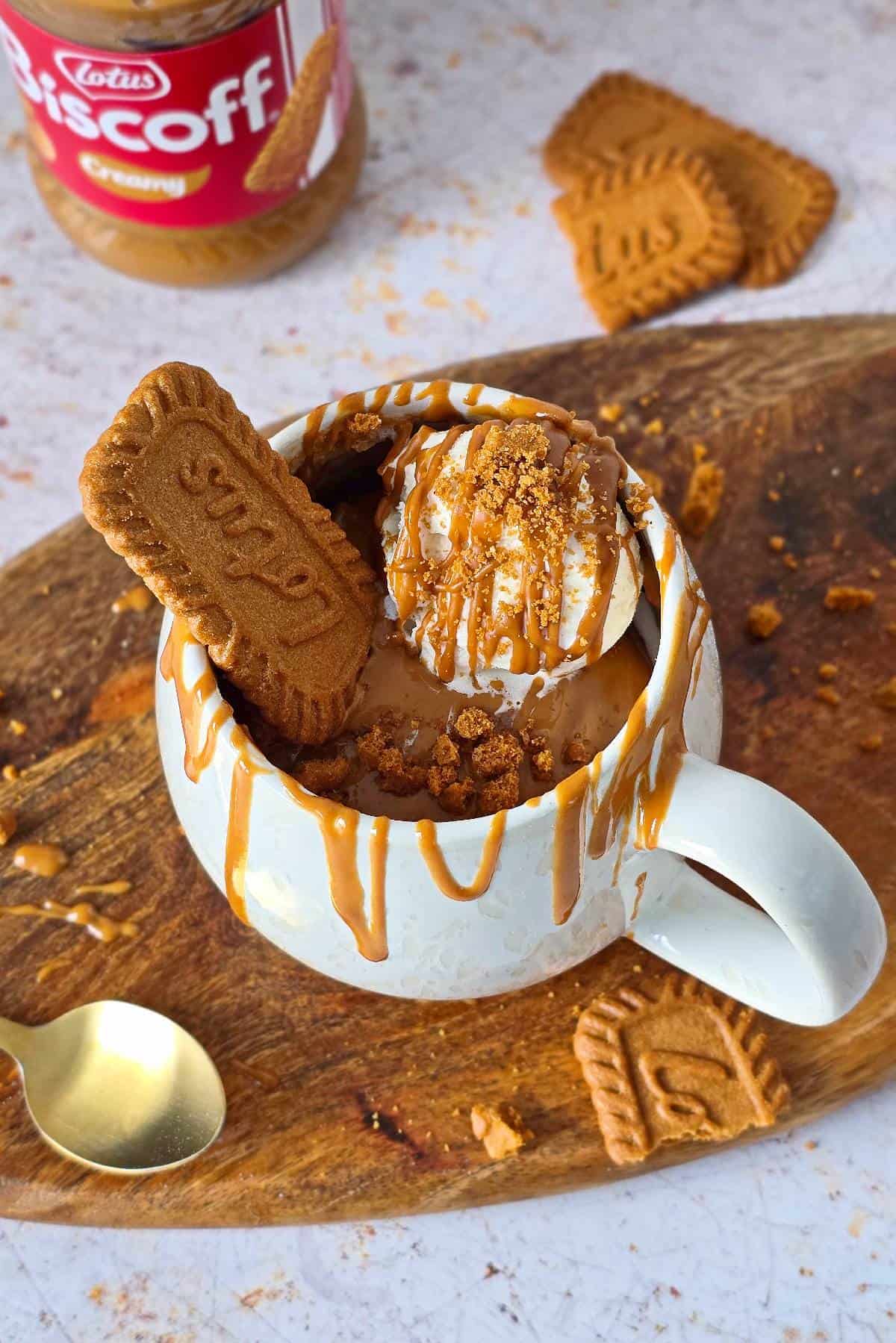 Biscoff mug cake in a white mug with ice cream, Biscoff drizzle, and cookies on a wooden platter, and gold spoon on the side.