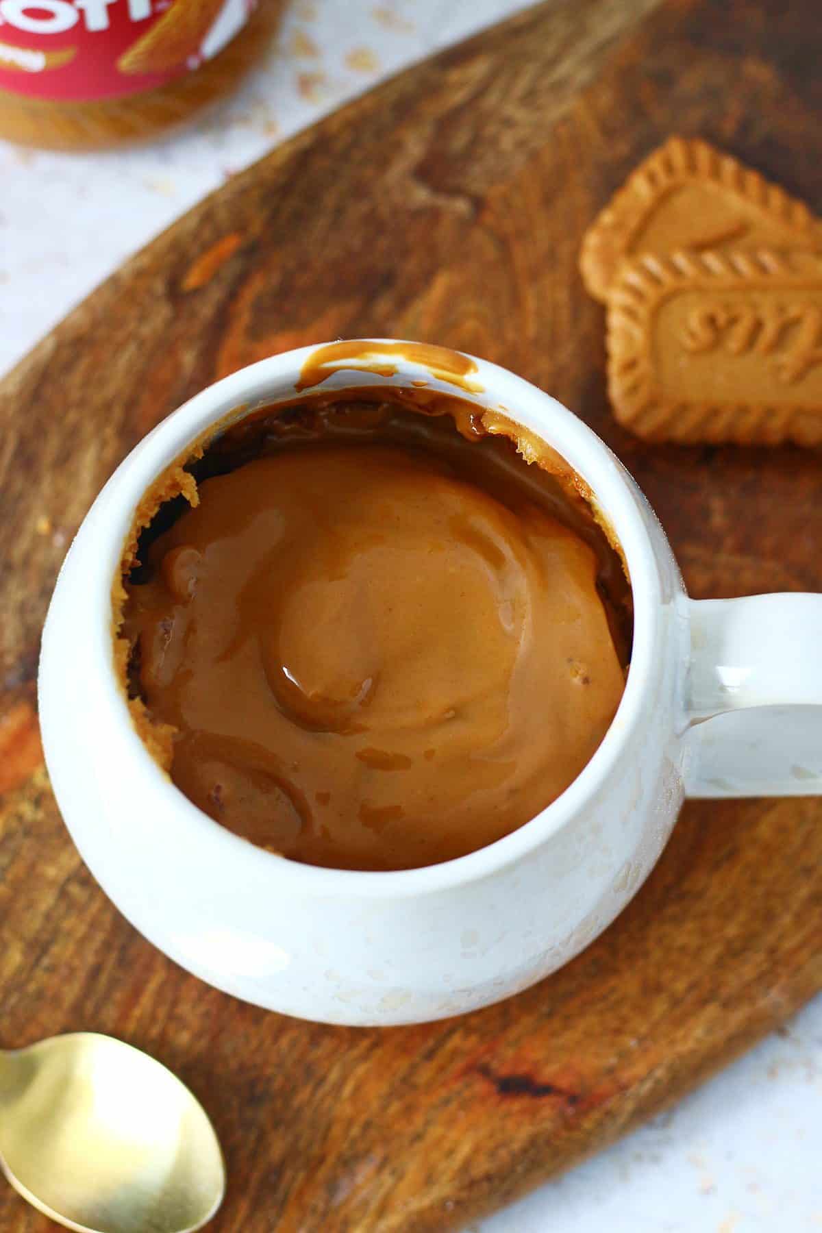 Biscoff mug cake with a glossy melted Biscoff topping in a white mug on a wooden surface.