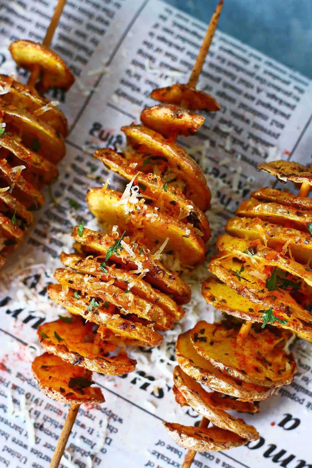 Close-up of tornado potatoes on skewers with crispy edges, seasoning, herbs, and grated parmesan on newspaper-lined surface.