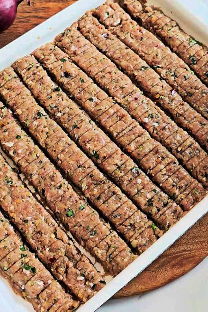 Raw kebab mixture shaped into strips and scored with lines on a baking tray.