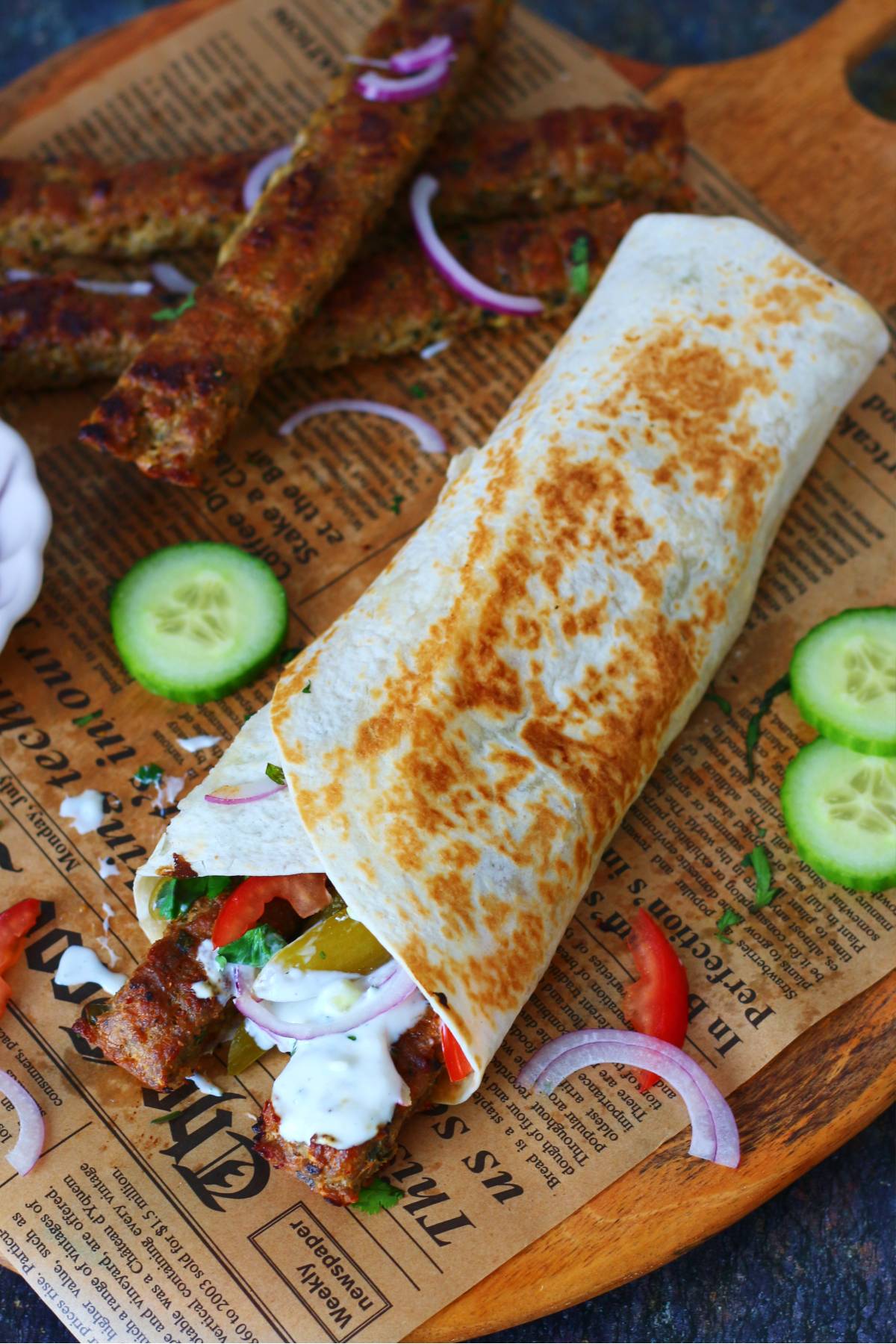 Kebab wrap filled with sheet pan kebabs, yogurt sauce, tomatoes, onions, and pickles on a wooden board.