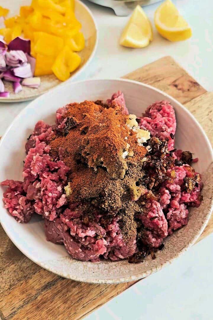 Ground beef with spices, fried onions, and aromatics in a bowl with chopped bell peppers and onions in the background.