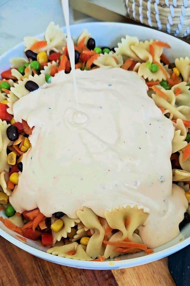 Creamy dressing being poured over bow tie pasta salad with vegetables and black beans in a large bowl.