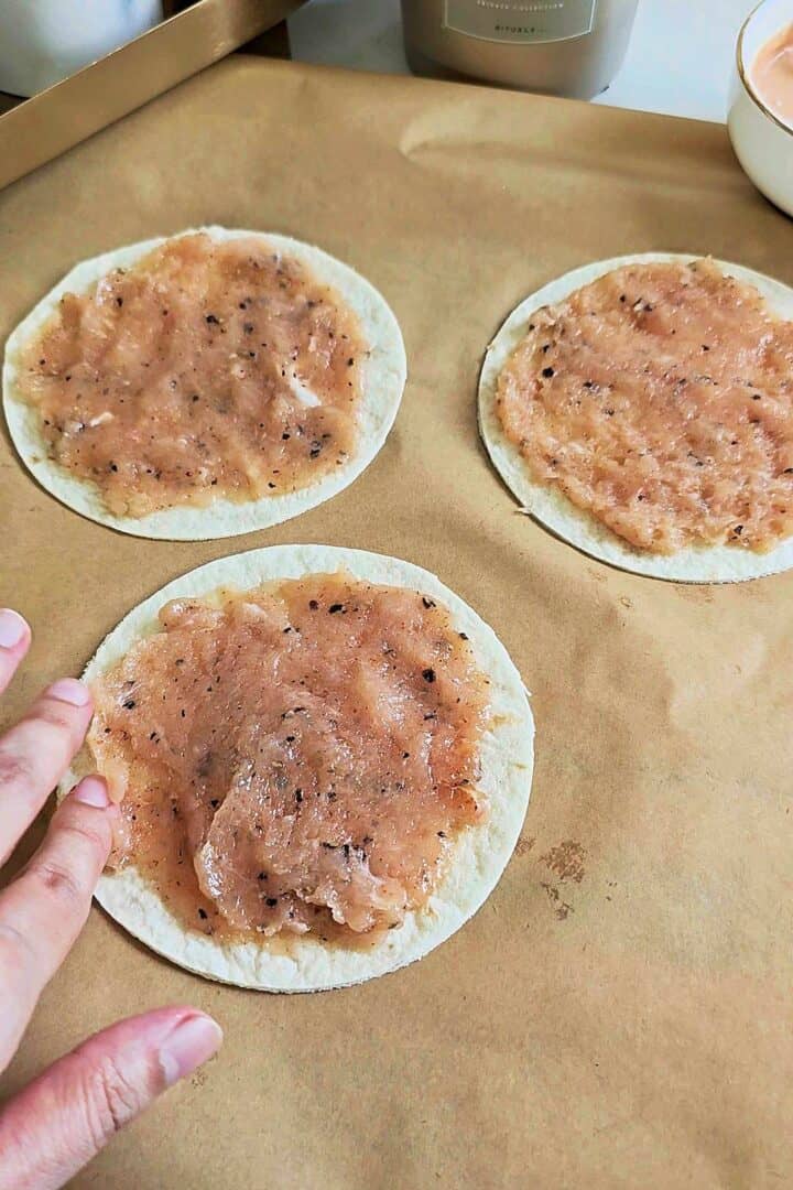 Mini flour tortillas topped with a thin layer of seasoned ground chicken on parchment paper.