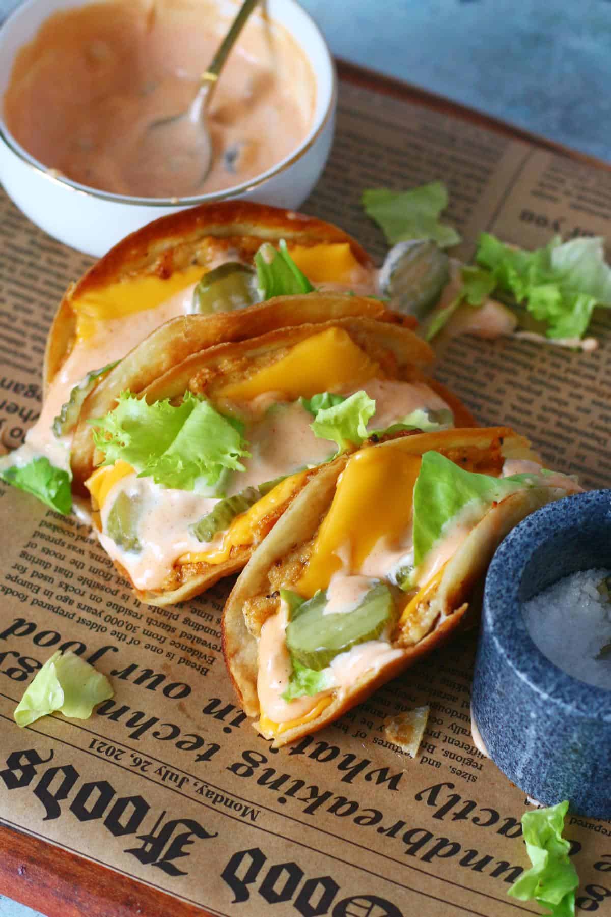Chicken Big Mac smash tacos topped with melted cheese, lettuce, pickles, and creamy sauce on parchment paper with a bowl of sauce in the background.