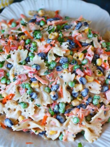 Creamy bow tie pasta salad served on a white plate with black beans, peas, sweet corn, carrots, and bell peppers.