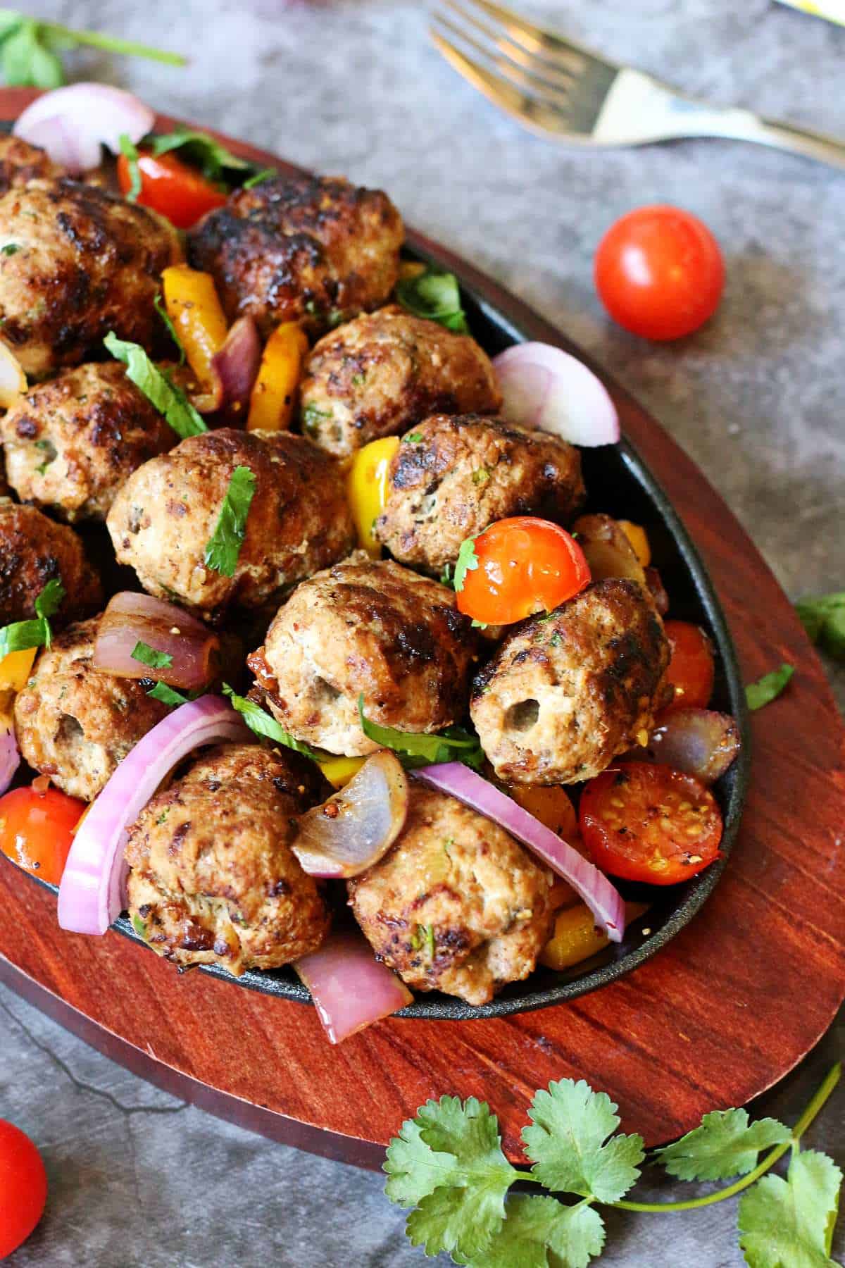 Close-up of browned gola kababs served with cherry tomatoes, onions, and bell peppers in a skillet.