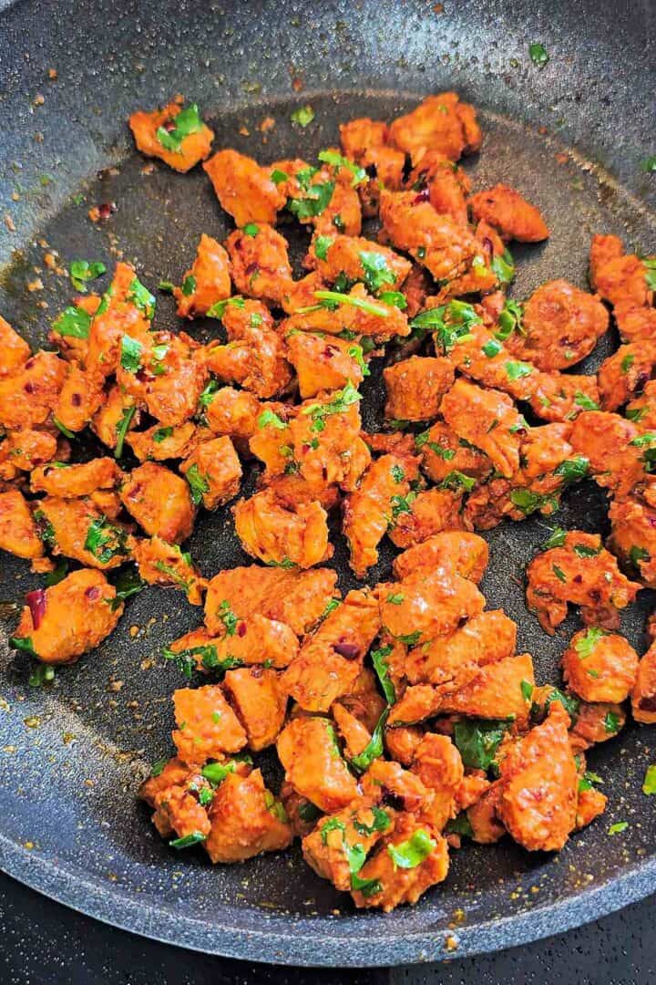 Cooked tikka chicken pieces tossed with fresh cilantro in a dark pan.