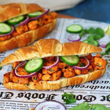 Two chicken tikka croissants on newspaper paper surrounded by fresh cilantro, cucumber and red onion.