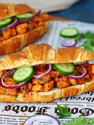 Two chicken tikka croissants on newspaper paper surrounded by fresh cilantro, cucumber and red onion.