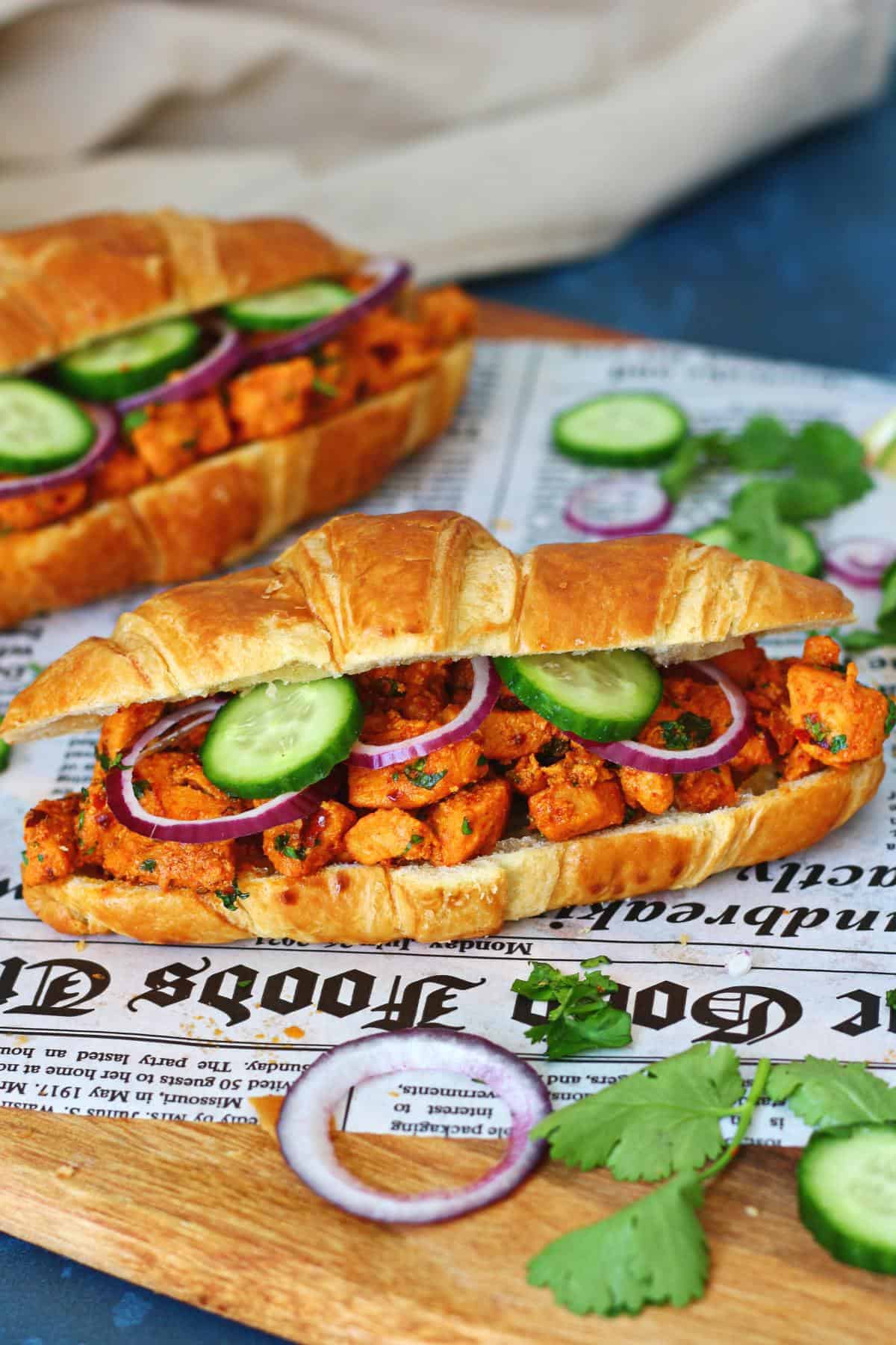 Two chicken tikka croissants on a wooden board with newspaper paper, garnished with cilantro, cucumber and red onion.