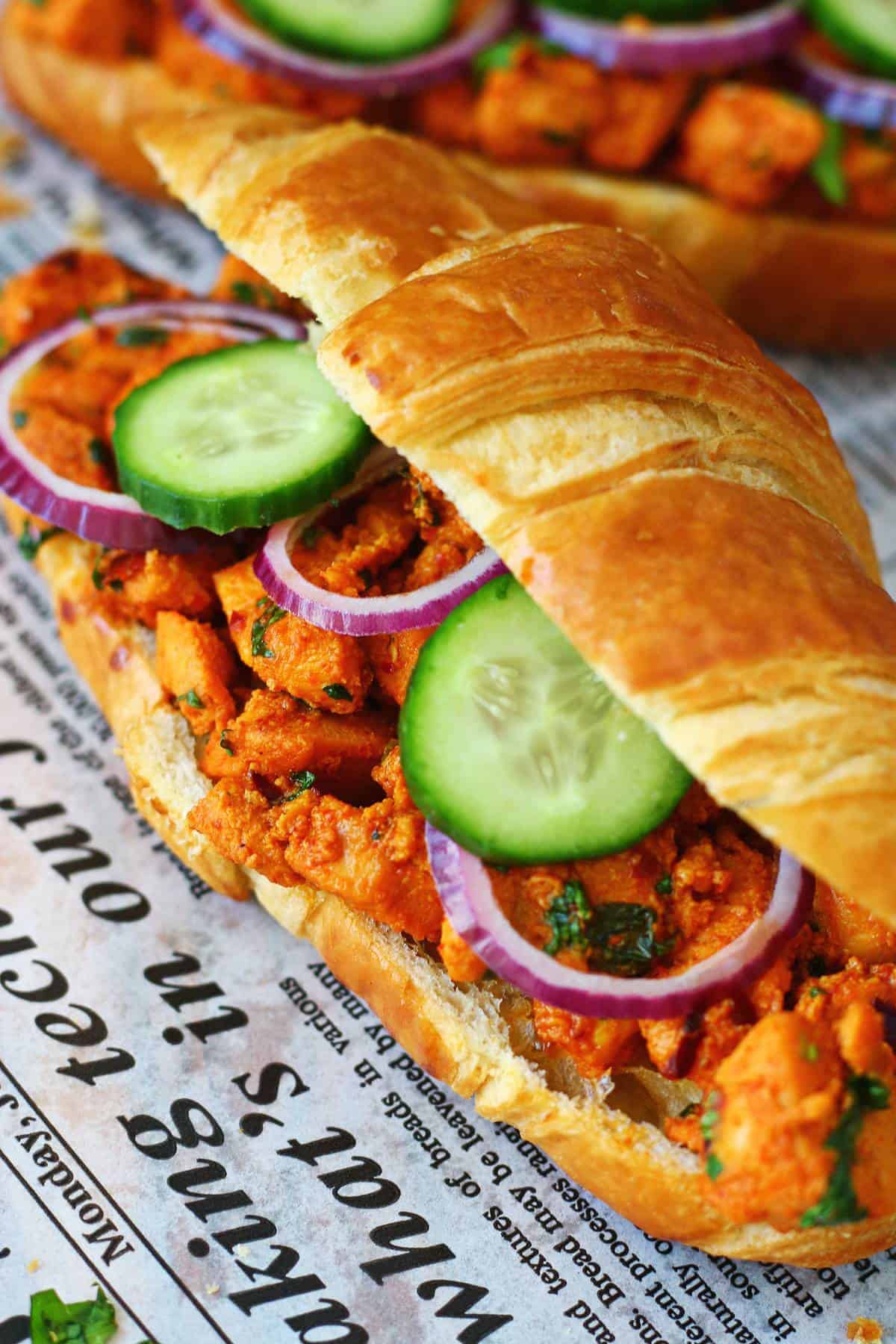 Side close-up of a chicken tikka croissant overflowing with spiced chicken, cucumber slices and red onion rings.