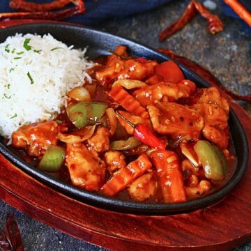 Dragon chicken served in a cast iron sizzler plate with white rice, bell peppers, onions, crinkle-cut carrots, and a glossy spicy sauce.