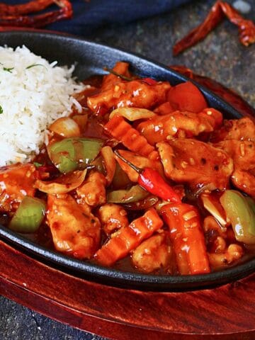 Dragon chicken served in a cast iron sizzler plate with white rice, bell peppers, onions, crinkle-cut carrots, and a glossy spicy sauce.