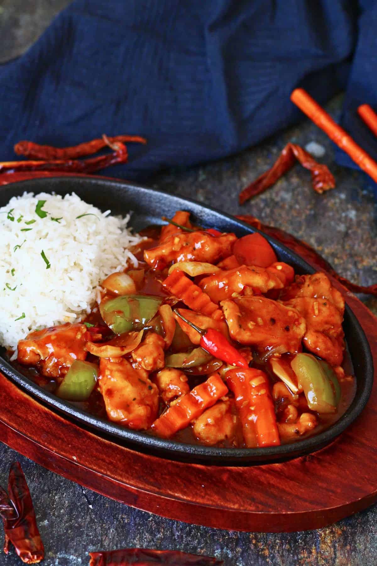 Dragon chicken in a cast iron sizzler plate with white rice, bell peppers, onions, crinkle-cut carrots, and dried red chilies scattered on a dark surface.