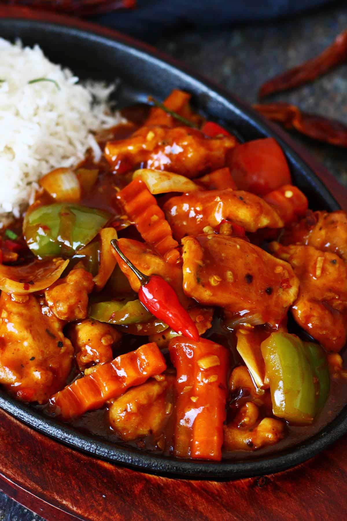 Dragon chicken in a cast iron sizzler plate with white rice, bell peppers, onions, crinkle-cut carrots, and a whole red bird's eye chili in a glossy spicy sauce.