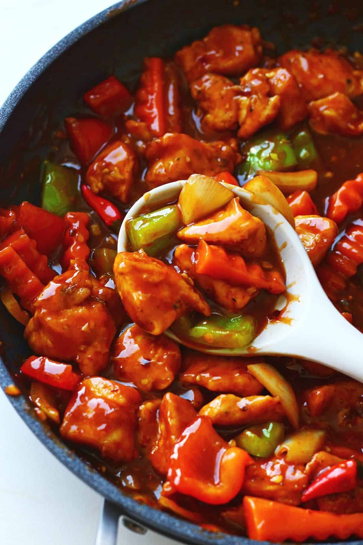 White spoon lifting dragon chicken coated in glossy spicy sauce with bell peppers, onions, and crinkle-cut carrots from a skillet.