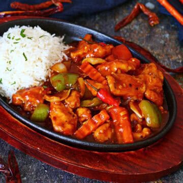 Dragon chicken served in a cast iron sizzler plate with white rice, bell peppers, onions, crinkle-cut carrots, and a glossy spicy sauce.