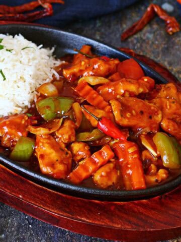 Dragon chicken served in a cast iron sizzler plate with white rice, bell peppers, onions, crinkle-cut carrots, and a glossy spicy sauce.