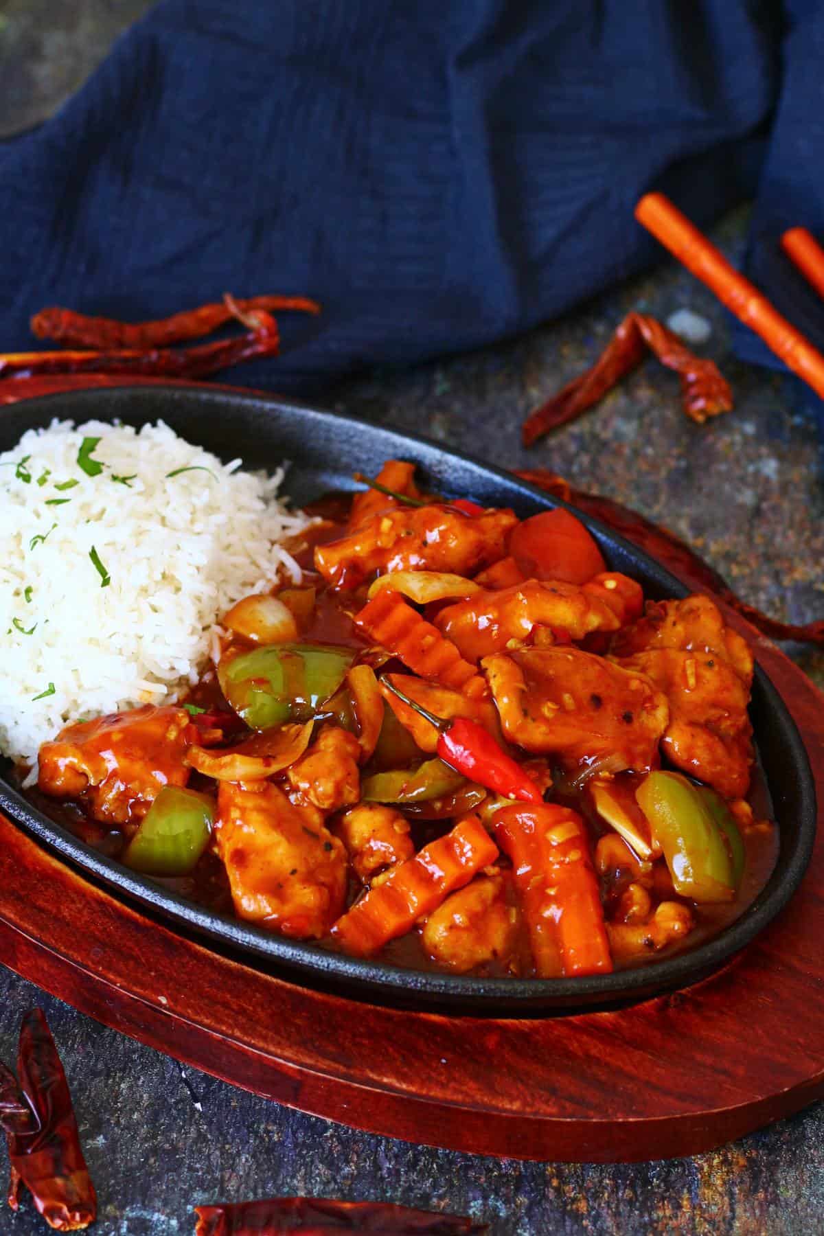 Dragon chicken in a cast iron sizzler plate with white rice, bell peppers, onions, crinkle-cut carrots, and dried red chilies scattered on a dark surface.