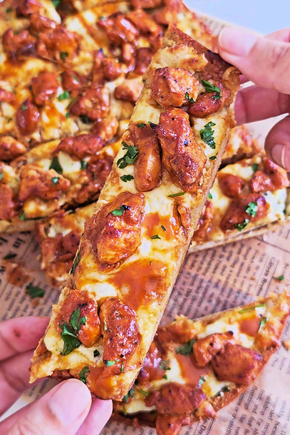 Hand holding a slice of buffalo chicken garlic bread topped with melted mozzarella, buffalo chicken, buffalo sauce, and chopped parsley.
