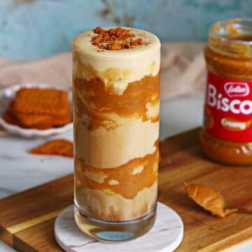 Iced Biscoff latte with cold foam in a tall glass, surrounded by Biscoff cookies and spread jar on a wooden board.