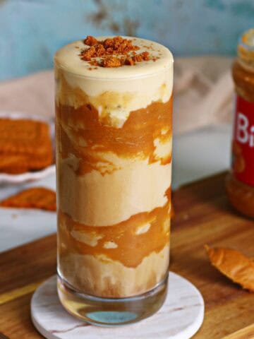 Iced Biscoff latte with cold foam in a tall glass, surrounded by Biscoff cookies and spread jar on a wooden board.