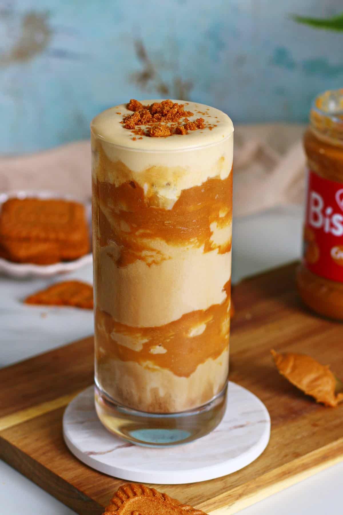Iced Biscoff latte with cold foam served in a tall glass with Biscoff spread jar and cookies on a rustic wooden board.