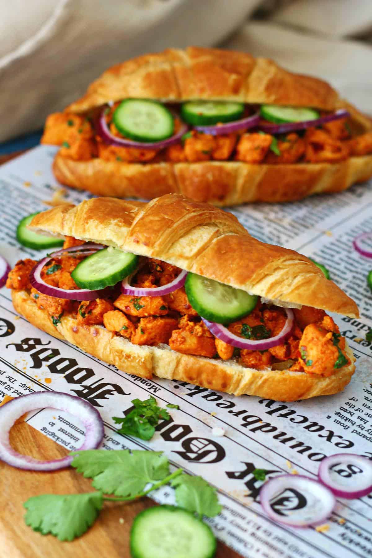 Two open chicken tikka croissants on a wooden board with cilantro, cucumber and red onion scattered around.