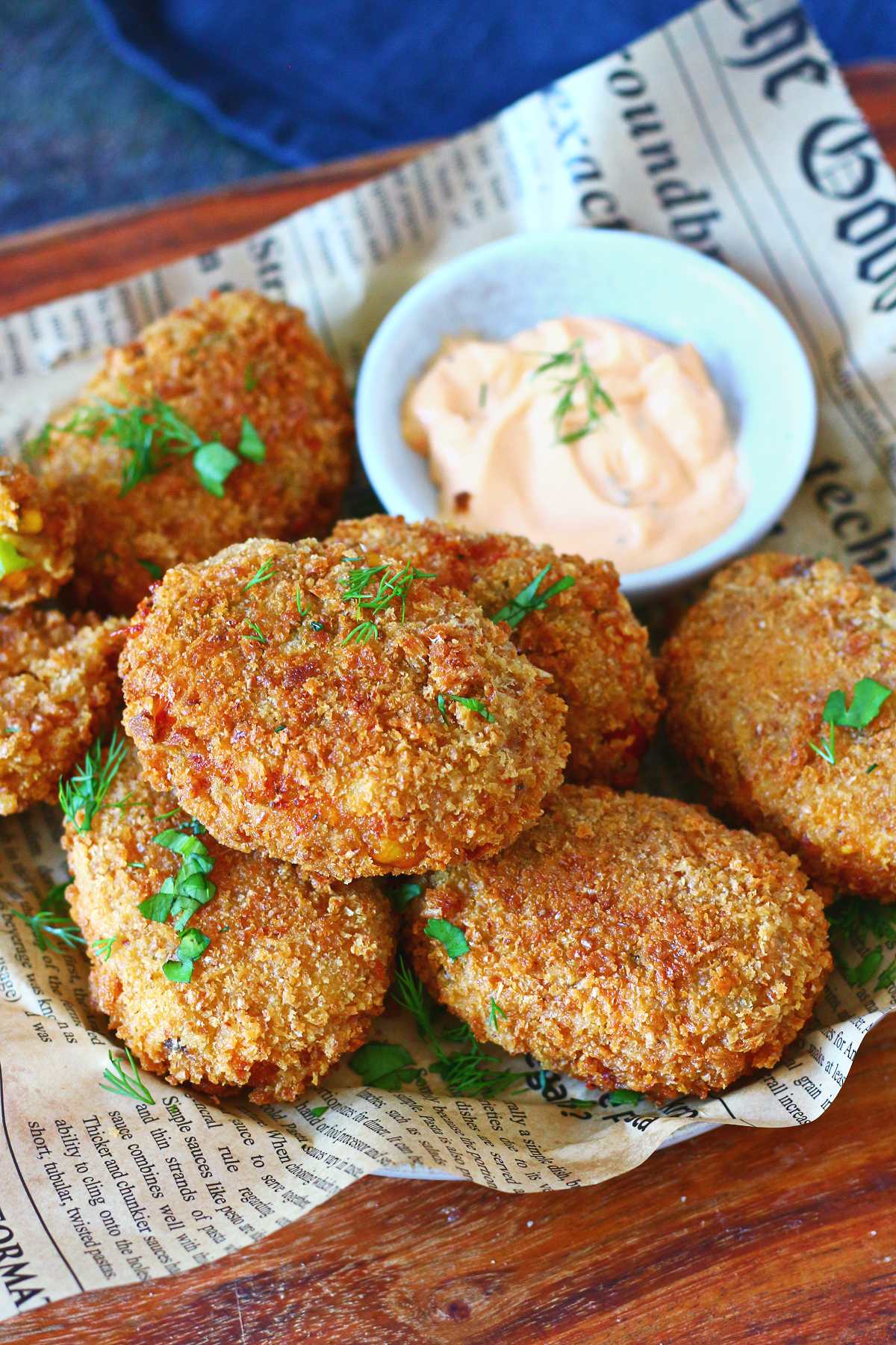 A stack of golden crispy cheesy chicken croquettes on newspaper paper, garnished with fresh herbs and served with a sriracha mayo dipping sauce.