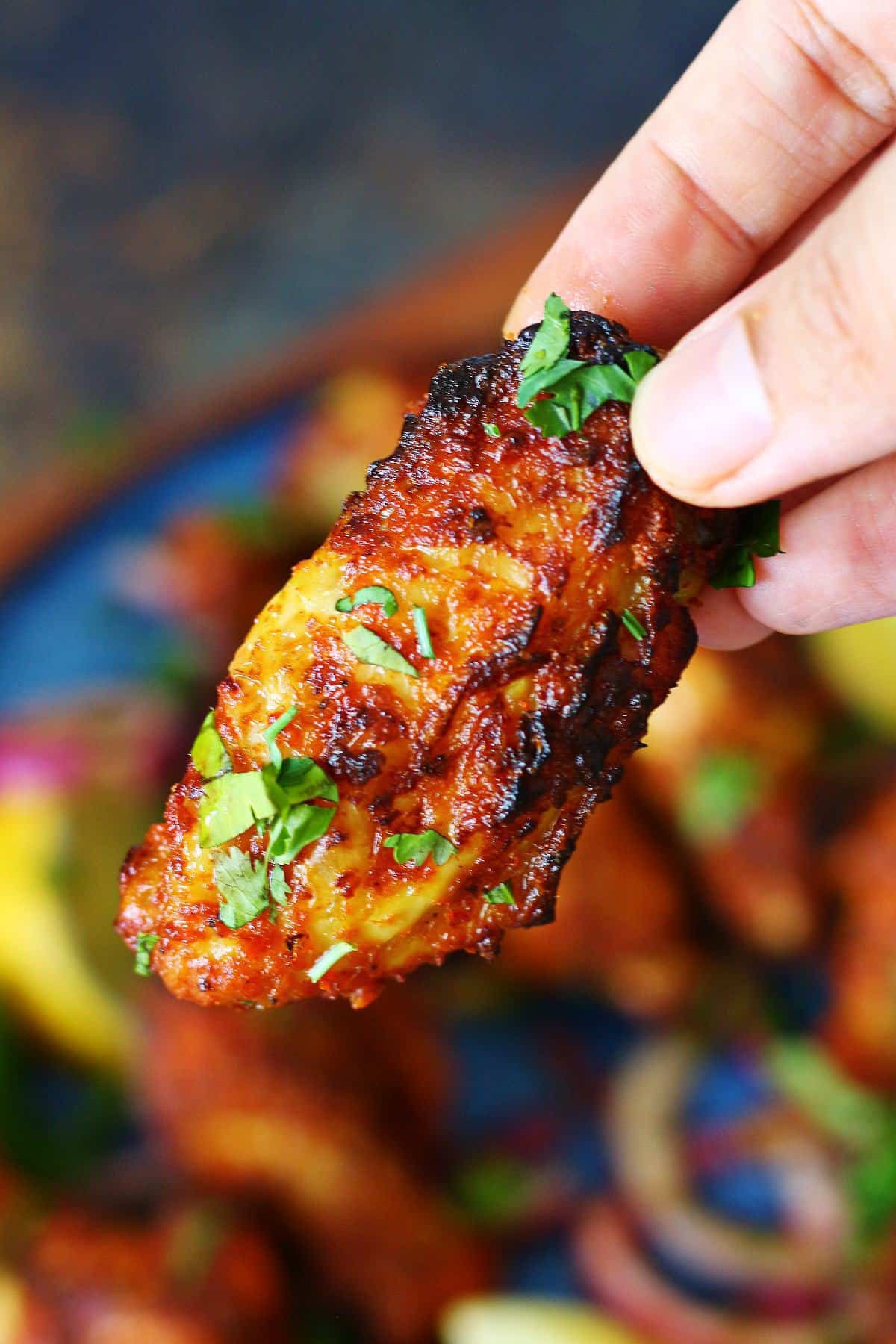Hand holding a charred air fryer tandoori chicken wing garnished with cilantro.