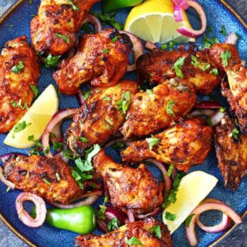 Overhead shot of tandoori chicken wings with charred skin, red onion rings, and lemon wedges.