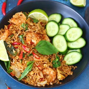Overhead view of Thai basil shrimp fried rice in a black bowl with cucumber slices and lime wedges.