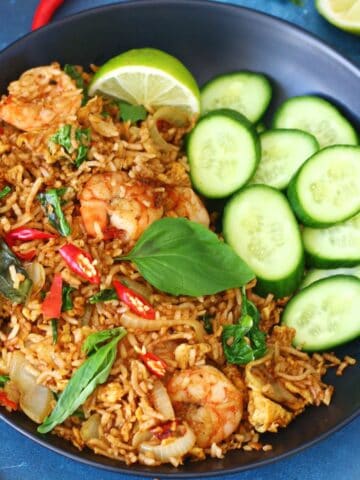 Overhead view of Thai basil shrimp fried rice in a black bowl with cucumber slices and lime wedges.