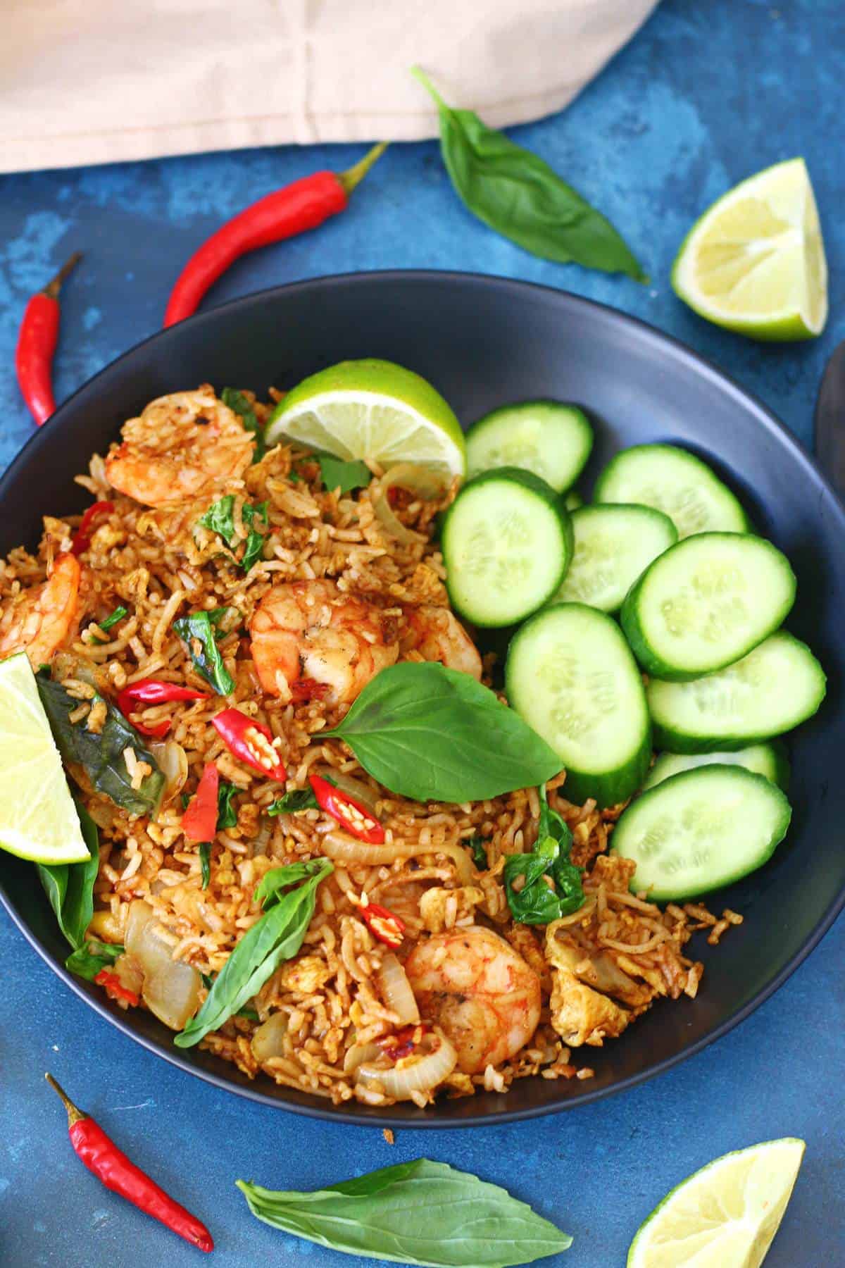 Thai basil shrimp fried rice in a black bowl, surrounded by bird's eye chilies, lime wedges, and fresh basil.