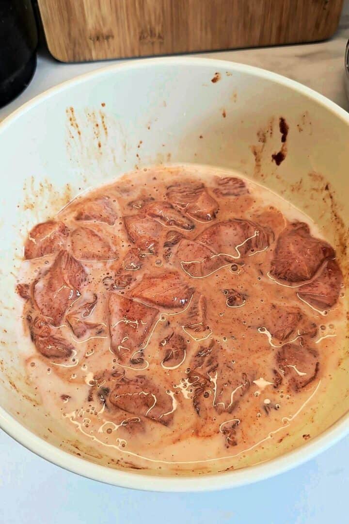 Bite-size chicken pieces soaking in a spiced buttermilk marinade in a white bowl.