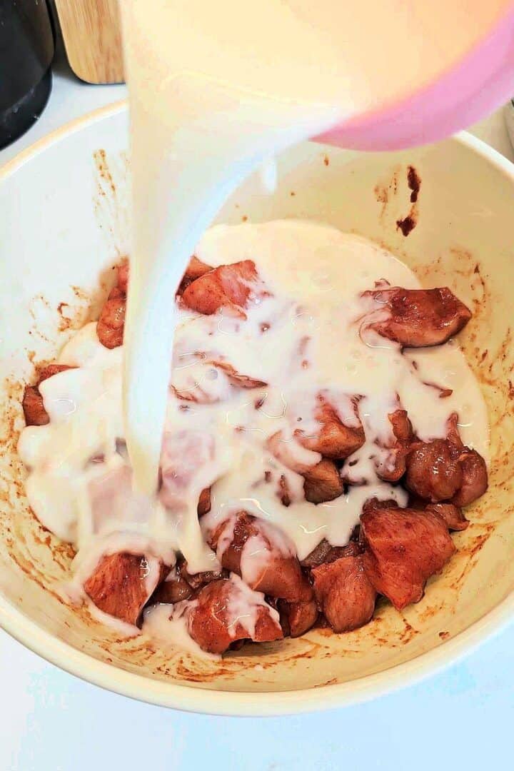 Buttermilk being poured over spiced chicken pieces in a white bowl.