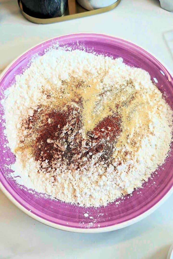 Flour, cornstarch and rice flour topped with cayenne, garlic powder and pepper on a purple plate, ready to be mixed.