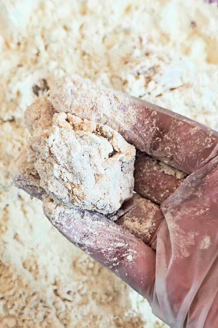 Gloved hand holding chicken pieces pressed in seasoned flour, showing the rough, craggy coating.